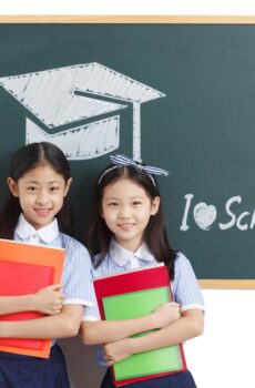 Two cheerful schoolgirls holding notebooks, standing by a chalkboard in a classroom.